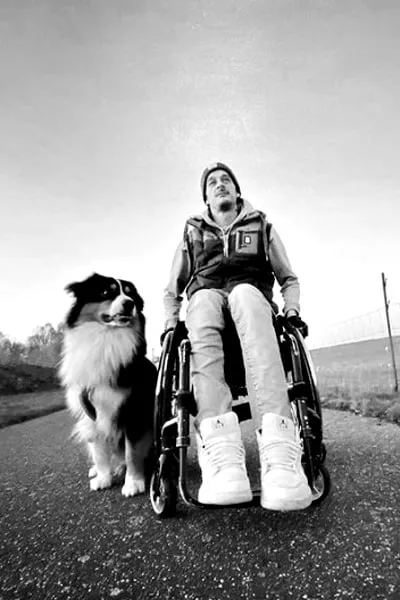 Christopher Calm sitzt im Rollstuhl auf einer Landstraße, neben ihm sitzt sein Hund. Schwarz-Weiß-Foto aus der Untersicht.